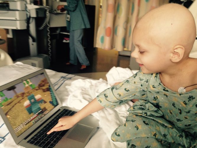 A child without any hair wearing a hospital gown pressing a computer keyboard while playing a game