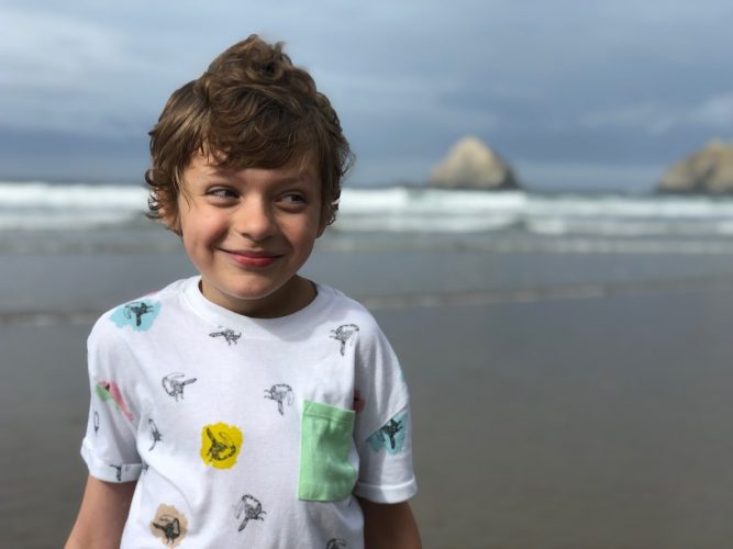 A young boy smiling while standing on a beach with large rocks in the waves behind him