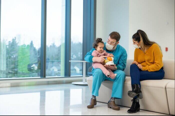 A little girl sits in her doctor's lap while her mom smiles and watches