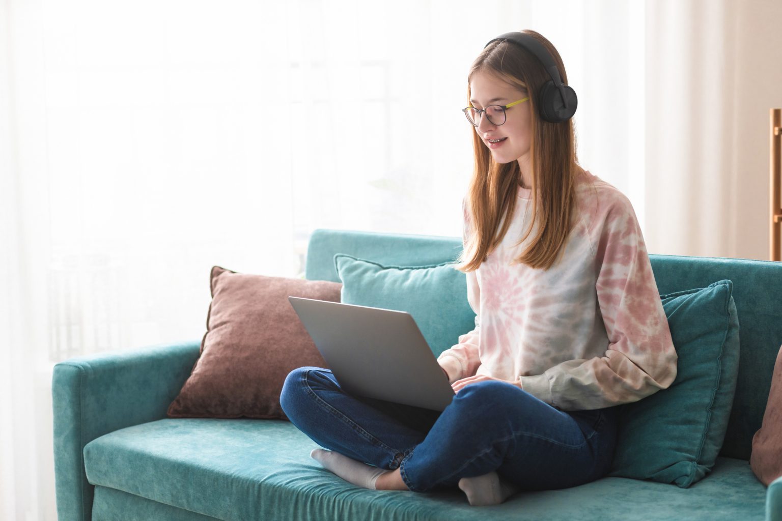 A teen sits on the couch wearing headphones while looking at a computer