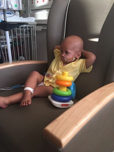 A toddler sits in a chair in his hospital room with his legs crossed and his hands behind his head