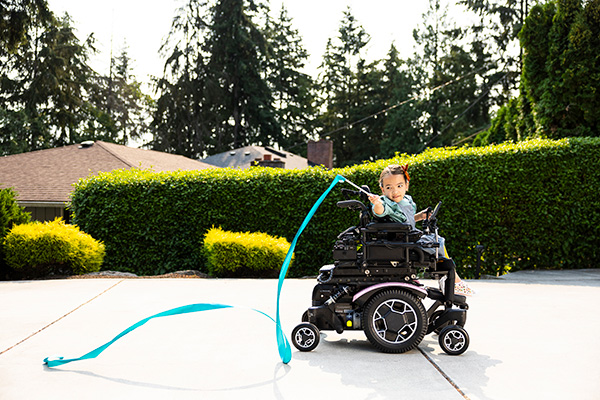 A young girl waves her teal dancing ribbon while she moves about in her motorized wheelchair