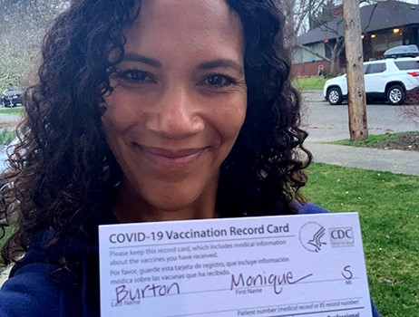 A doctor smiles at the camera as she holds up her vaccination card