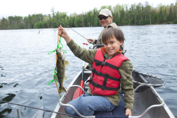 A boy in a red life jacket holds up a fish while sitting in a canoe on a lake with his dad