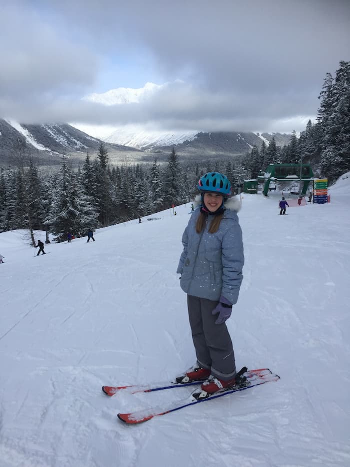 A young girl wearing winter gear and skis, smiles from the slope