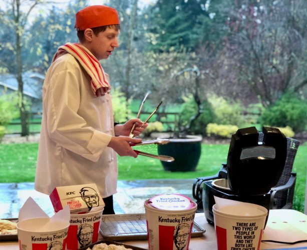 A young man wearing a white chef's jacket and holding two metal tongs stands near a fryer while the table is covered in KFC containers