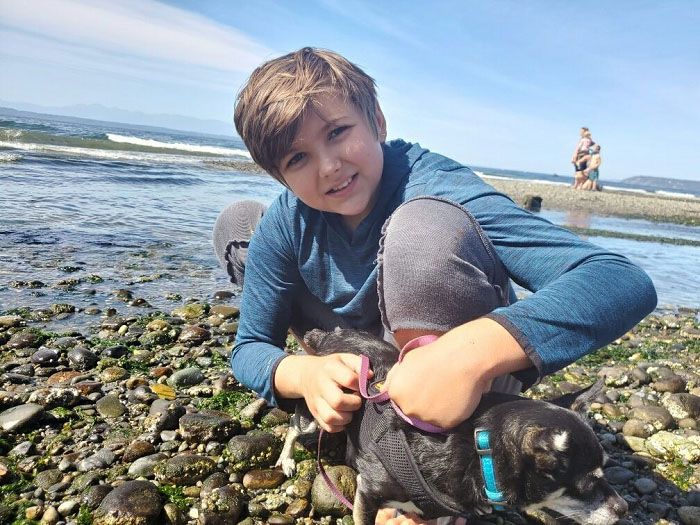 A young boy kneels down with his small dog while looking at tide pools on a beach