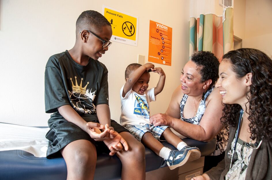 Two happy kids smile while sitting on a medical exam table while two women smile back at them