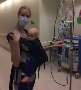 a mother carries her son in a Baby Bjorn carrier in the hospital while pulling his IV pole