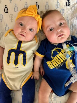 two twin preemies smile at the camera while wearing Seattle Mariners onezies