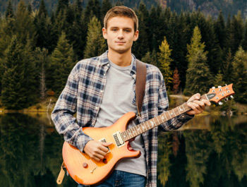 A teen boy stands with a guitar in front of a forest lake