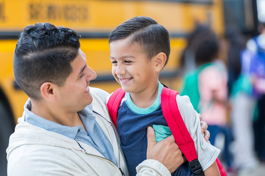 A closeup photo of a dad and son smiling at each other with a yellow school bus in the background