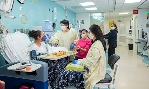 Mary sits in a hospital bed while a medical provider takes her temperature on her forehead. Her sister Monica is sitting at her bedside and another medical provider sits in a chair at the end of the bed.