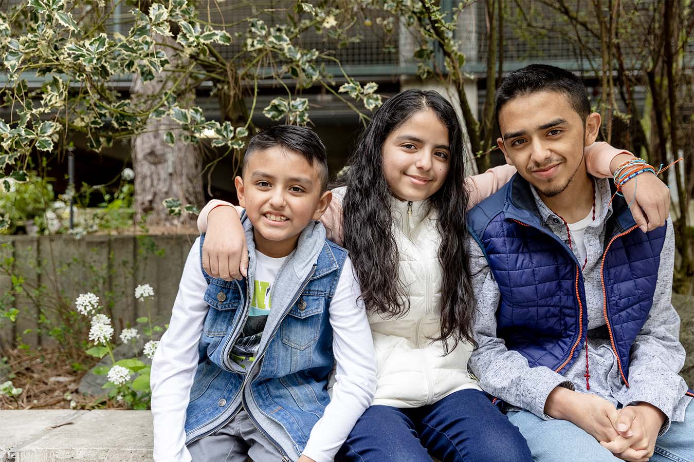 Two children and an adolescent sit together on a bench smiling at the camera