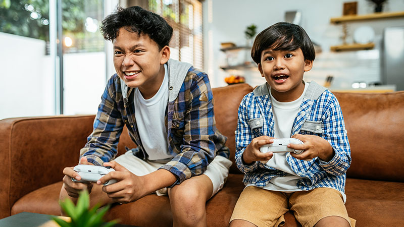 Two boys sit next to each other on a brown leather couch with game controllers in their hands and intense looks on their faces