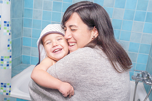 A woman smiles as she hugs her smiling baby who is wrapped in a white bath towel against a light blue tile background