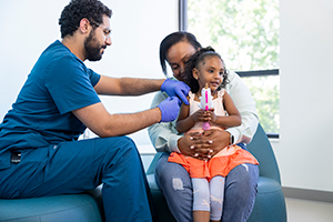 A young girls sits on her mom's lap while a male provider in blue scrubs gives her a vaccination